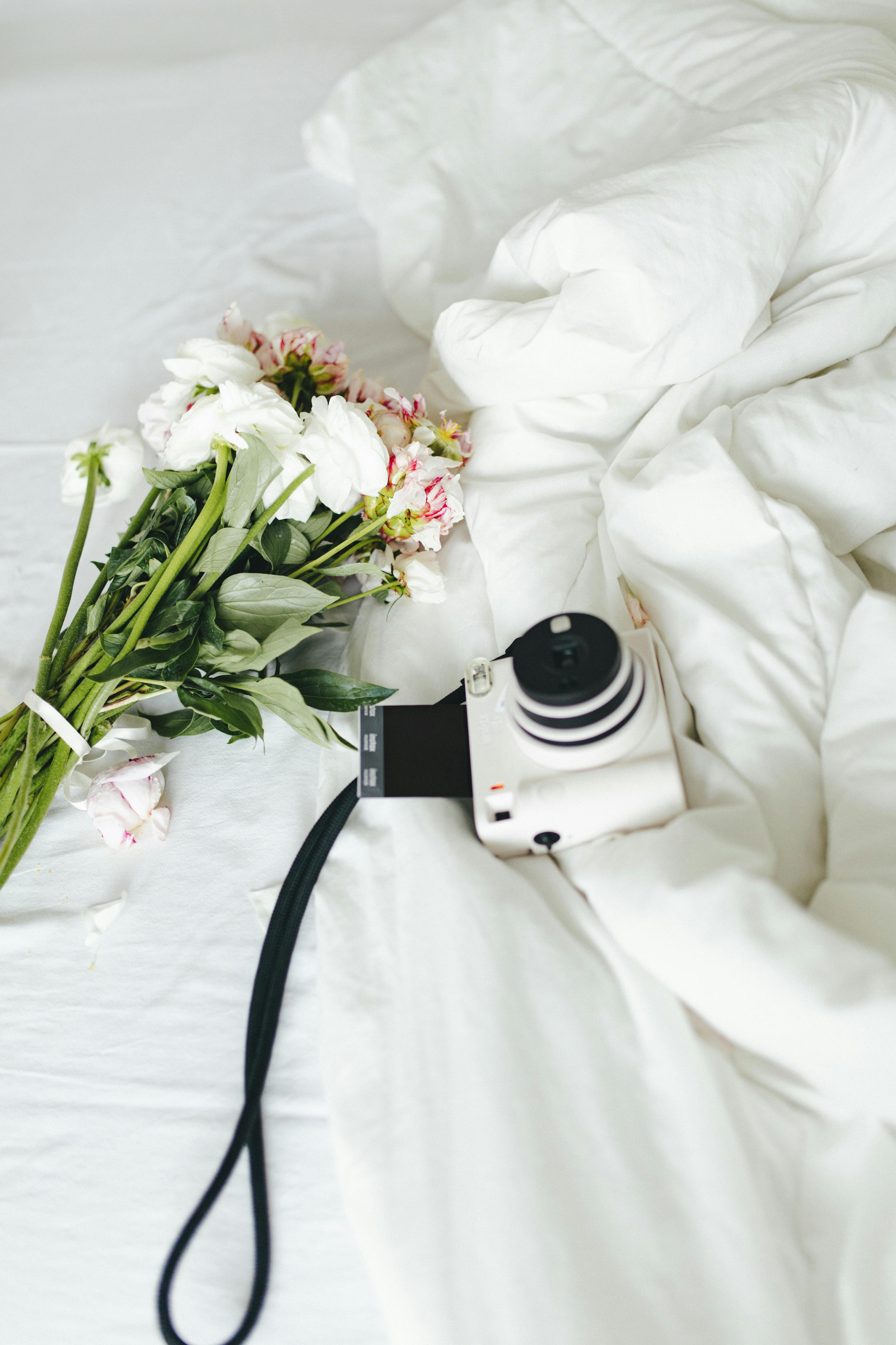 A white instant camera and fresh flowers on a cozy white bed. Perfect for lifestyle themes.