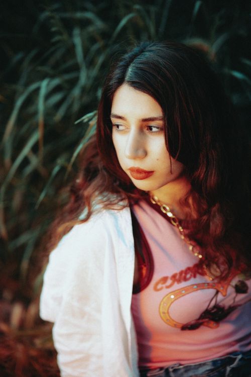 Free Young woman with long hair wearing a pink shirt outdoors, looking thoughtful adjacent to foliage. Stock Photo