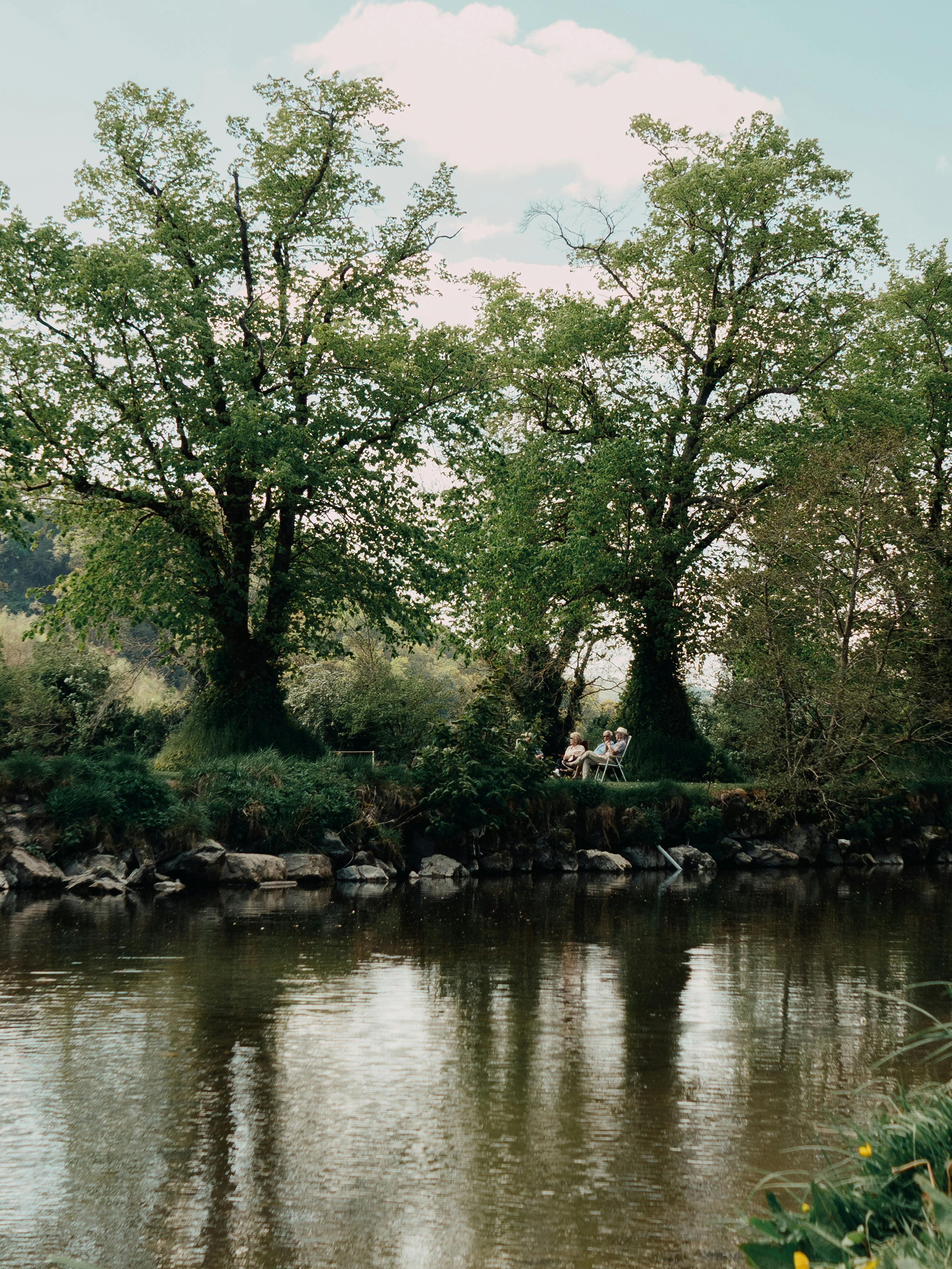 Serene scene by the river with two people relaxing under lush trees in Llanfair Talhaiarn, Wales.