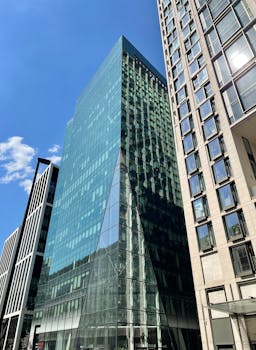 Tall glass skyscraper surrounded by urban office buildings under clear blue sky.
