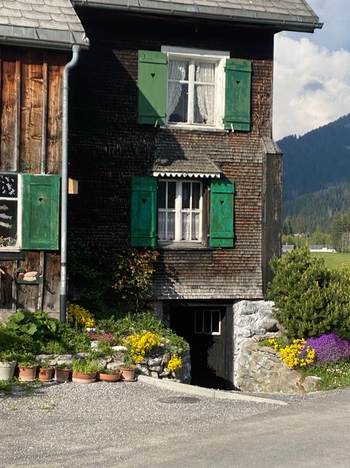 Free A rustic house with green shutters, surrounded by vibrant flowers, under a clear sky. Stock Photo
