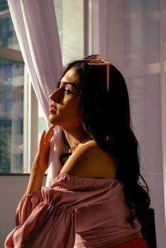 Portrait of a woman in a sunlit room, wearing a pink top and headband, gazing out the window.