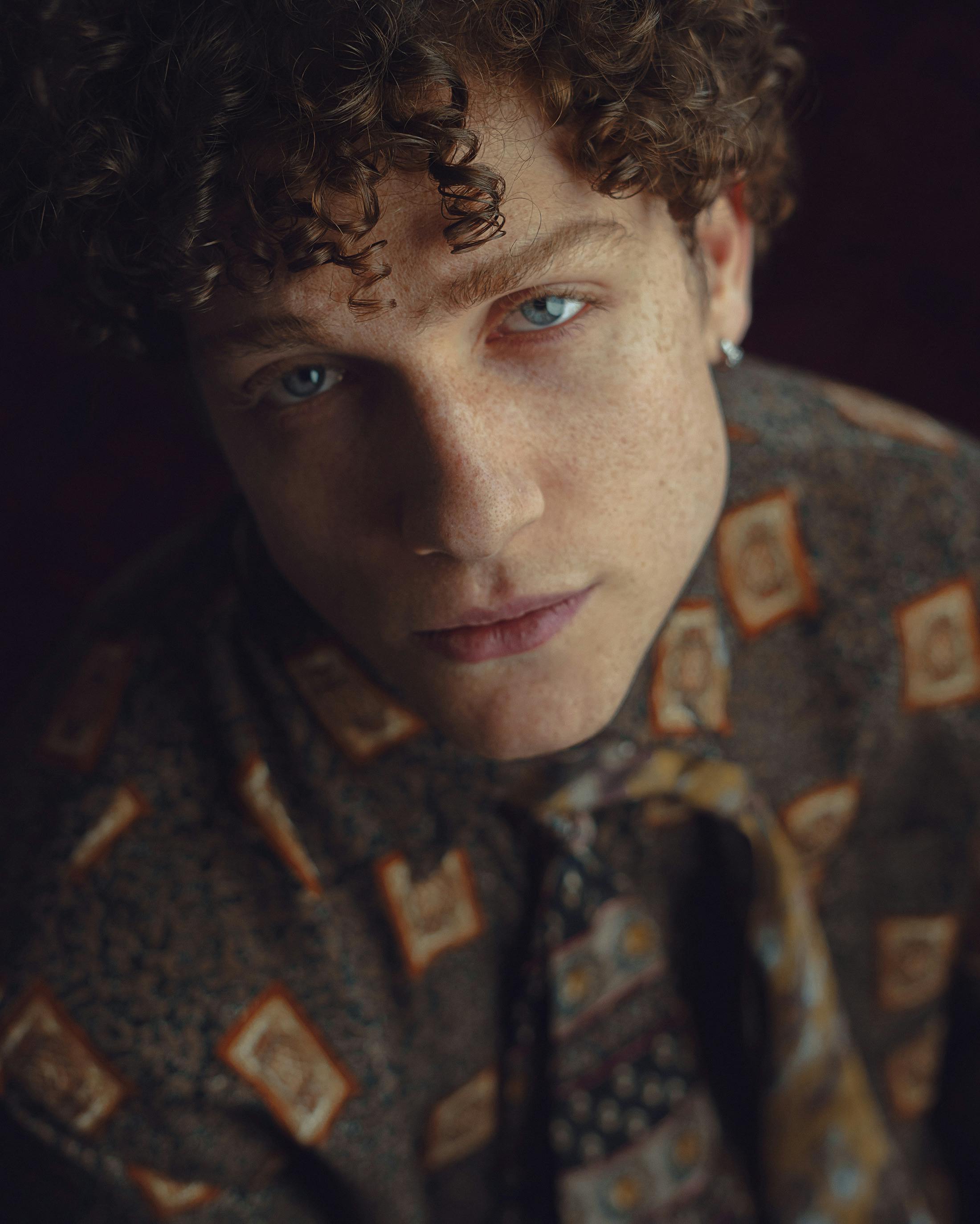 Portrait of a young man with curly hair and patterned vintage shirt, looking confidently at the camera.