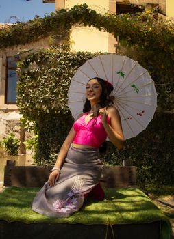 A woman poses gracefully in a vibrant outfit with a parasol in a sunny garden setting.
