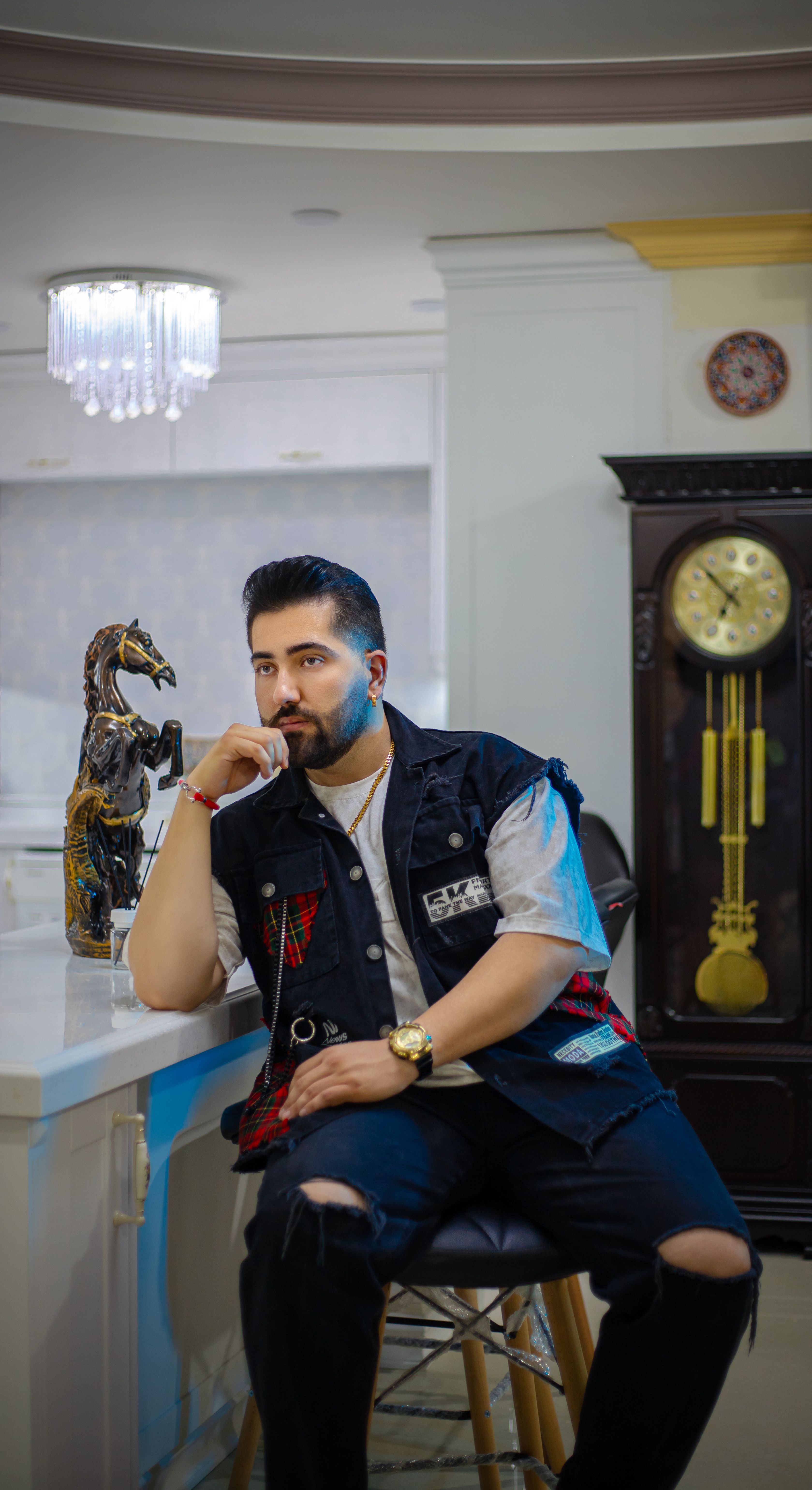 Man in casual attire poses in a modern setting with a vintage clock and decor, exuding confidence.