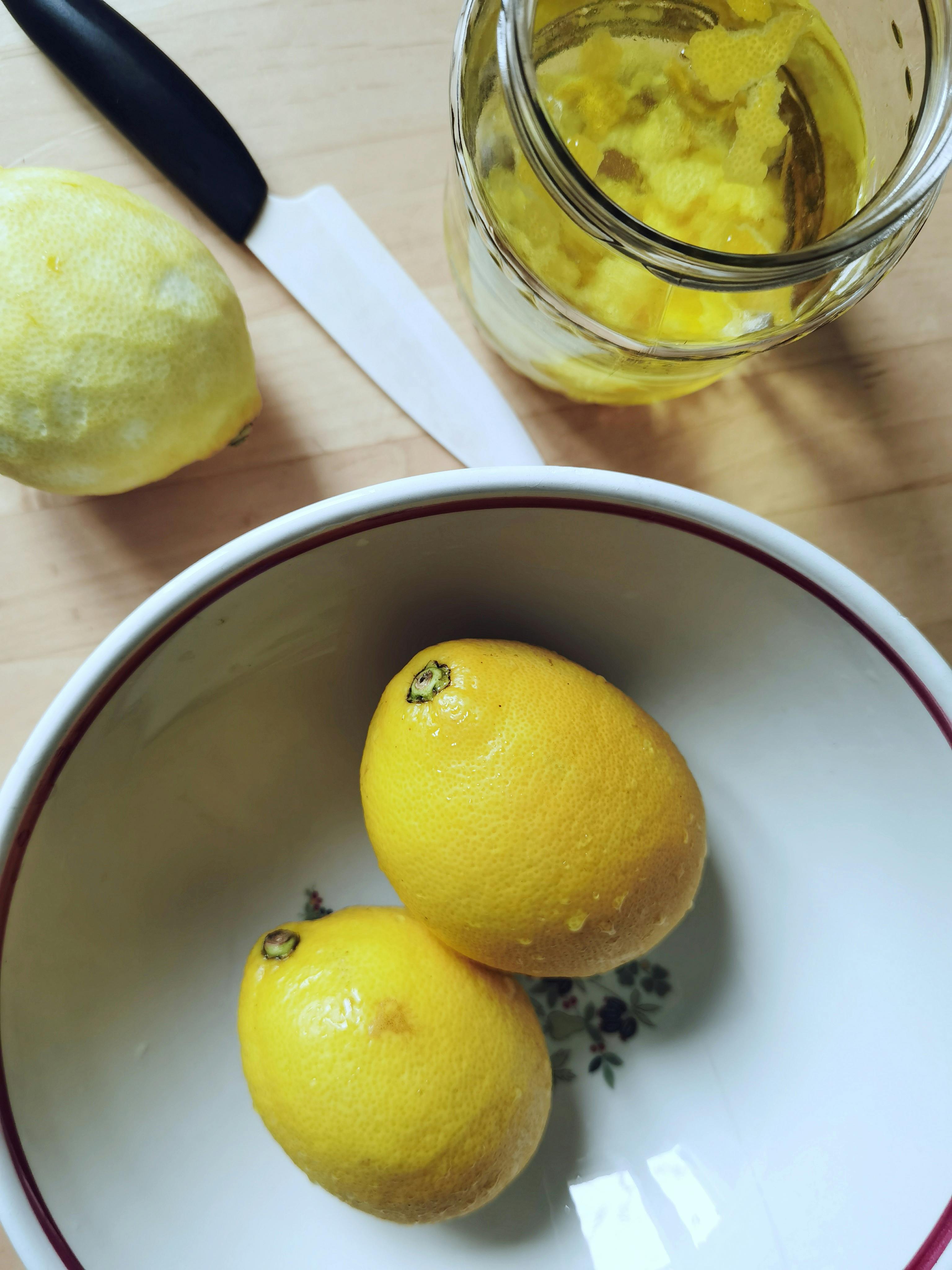 Top view of fresh lemons in a bowl with a jar of lemon preserve on a wooden surface.