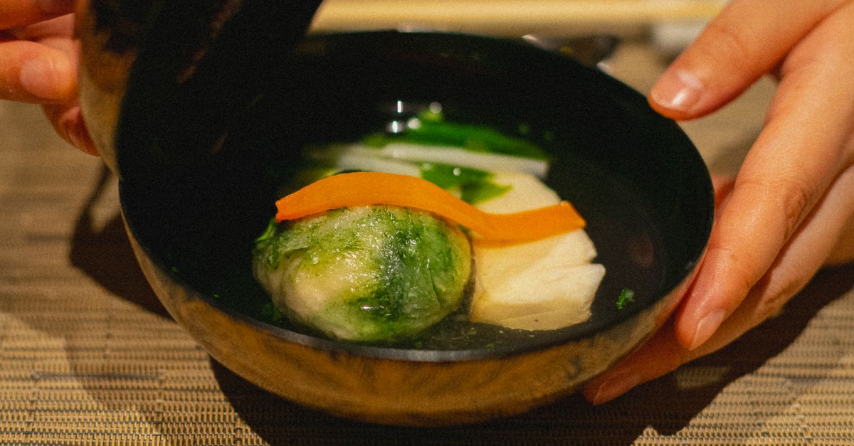 Hands holding a bowl of miso soup with steam rising, ingredients around it