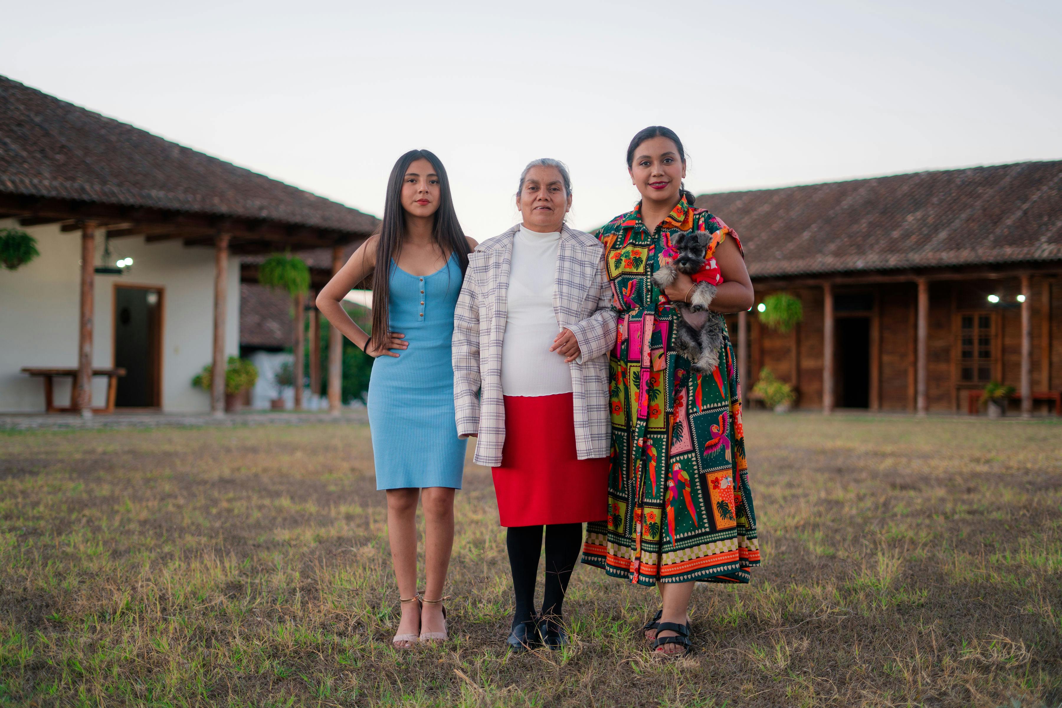 Three women stand together outside a traditional rustic house, showcasing diverse cultural attire.