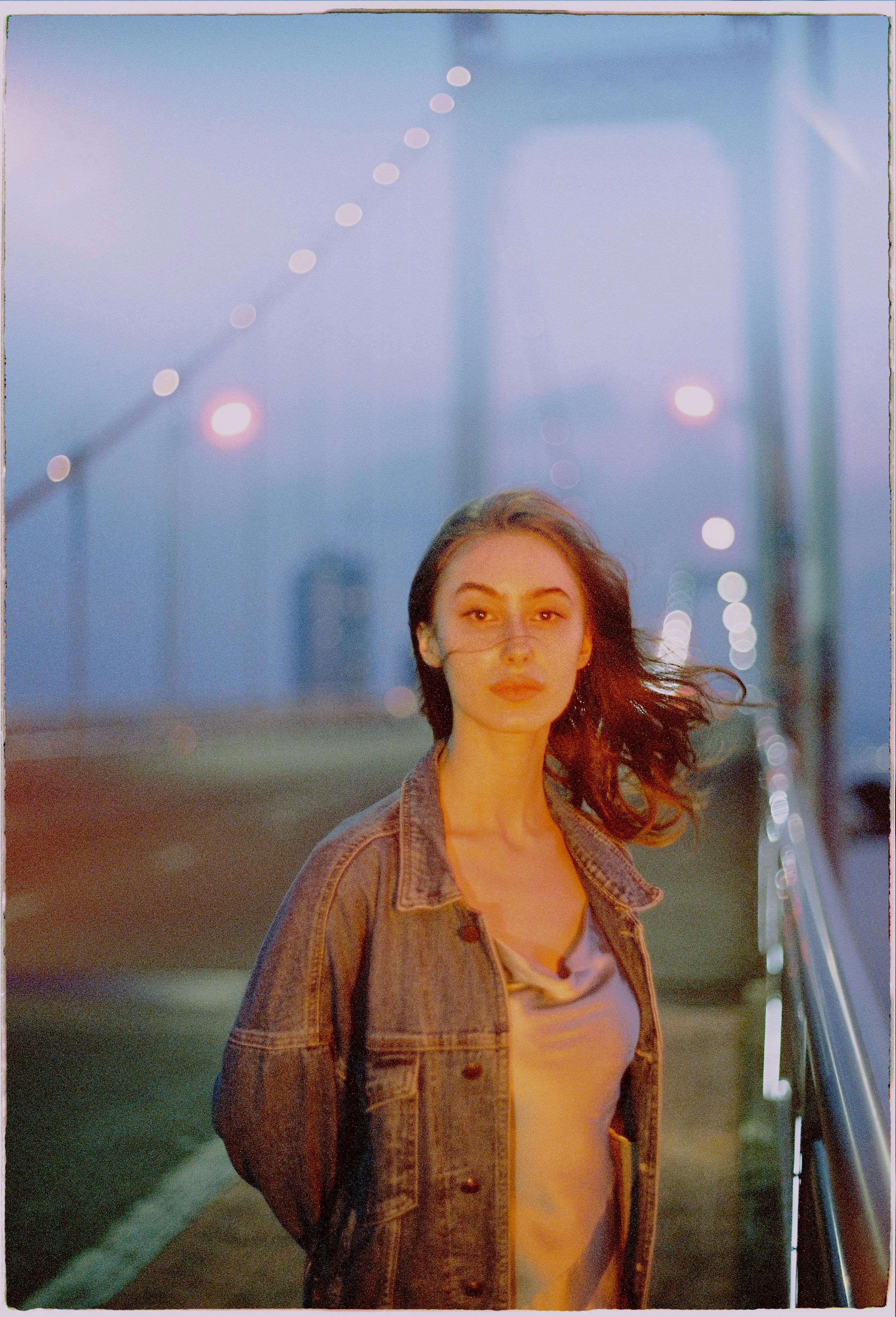 Young woman stands on a lit bridge at dusk, creating a serene urban portrait.