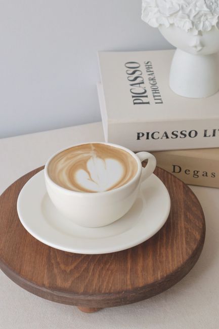A cup of espresso with latte art on a wooden tray beside art books in a cozy setting.