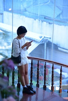 A young woman texting on smartphone by glass window indoors, wearing casual attire.
