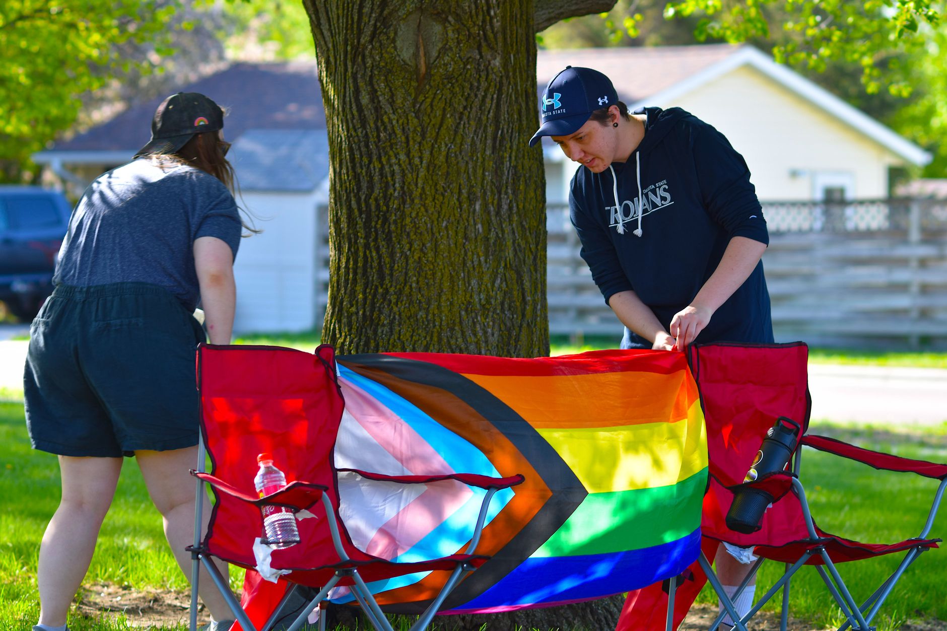 Two people organize a picnic with a Progress Pride flag under a tree.