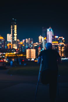 Senior man with crutches gazes at vibrant city skyline at night, reflecting solitude and urban life.