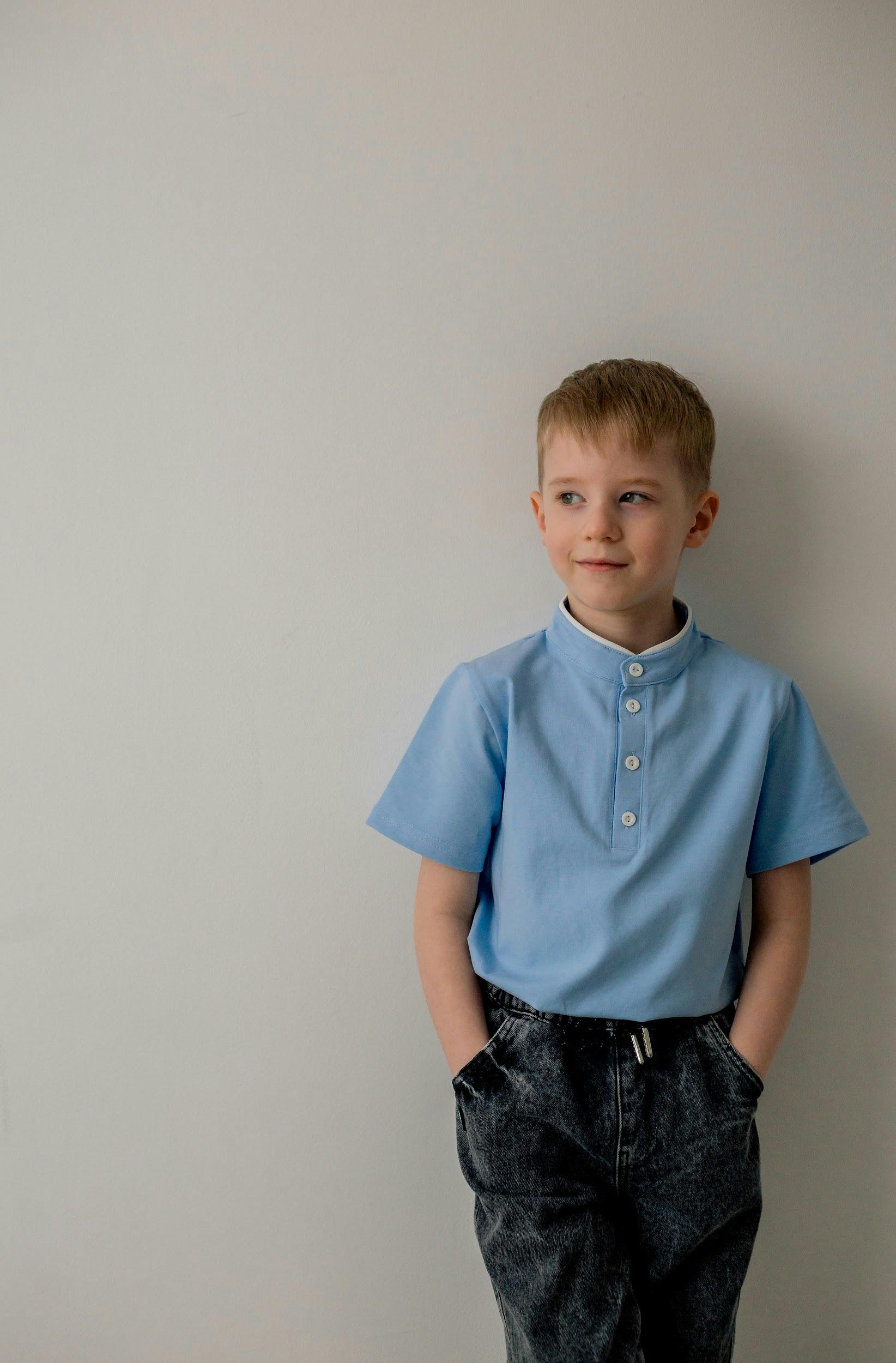 gratis Een jonge jongen in een blauw shirt en spijkerbroek staat tegen een lichte muur en kijkt nadenkend. Stockfoto