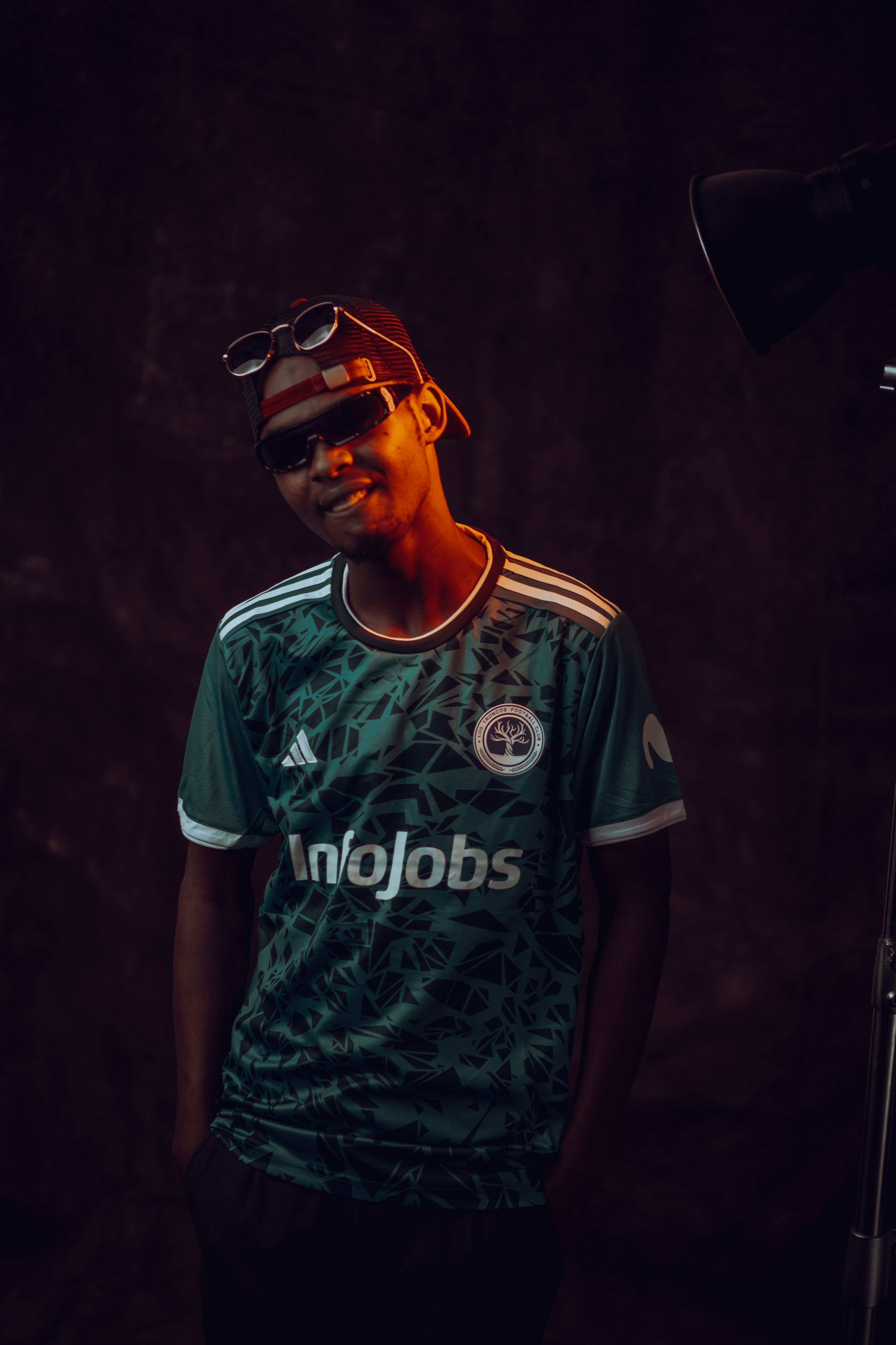 Cool young man wearing sunglasses and a green soccer jersey posing indoors.