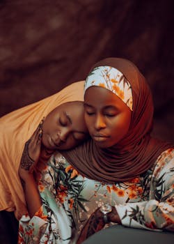 Two women wearing hijabs share a tranquil moment, with traditional attire and a warm, intimate setting.