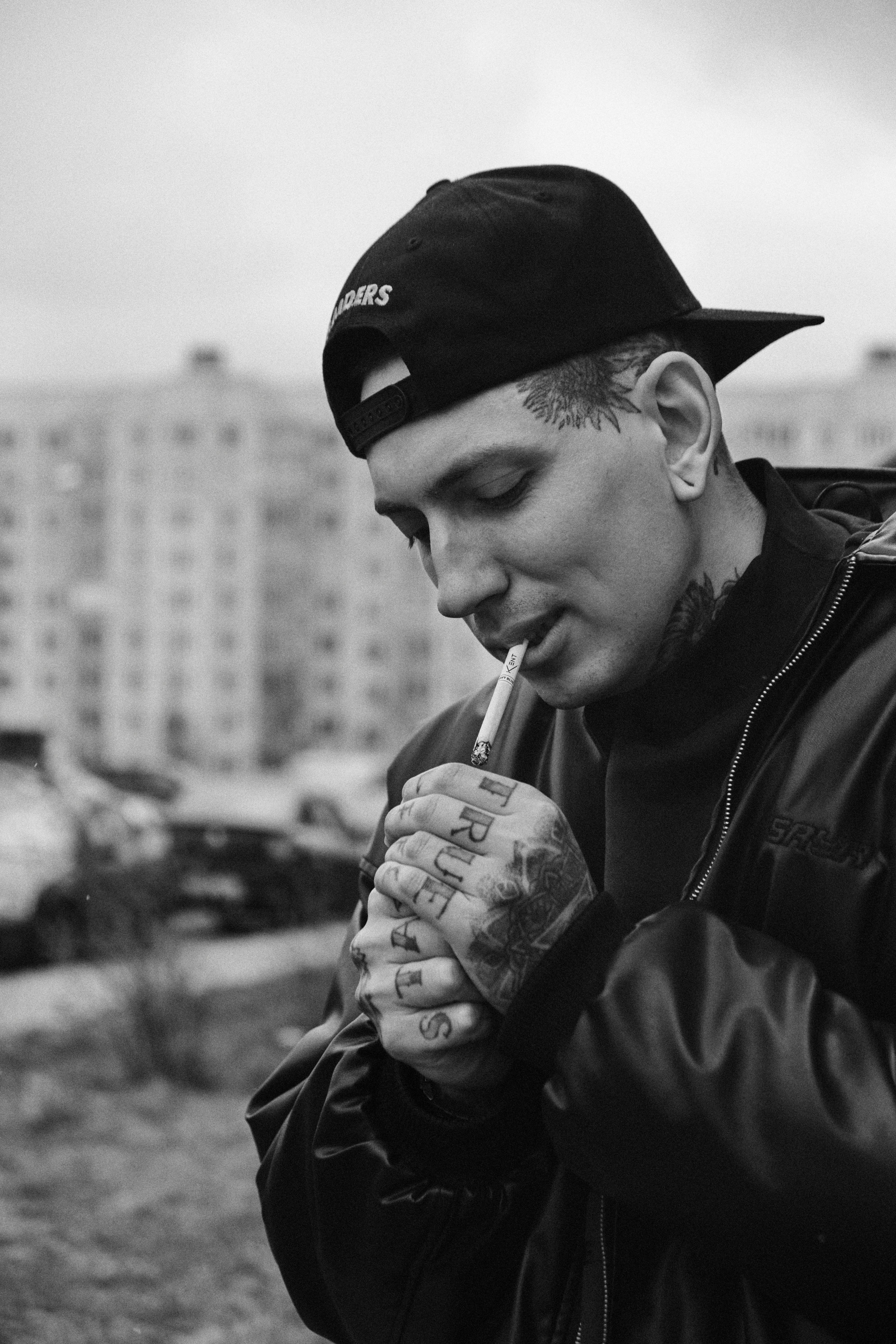 A tattooed man in a baseball cap lighting a cigarette outside.