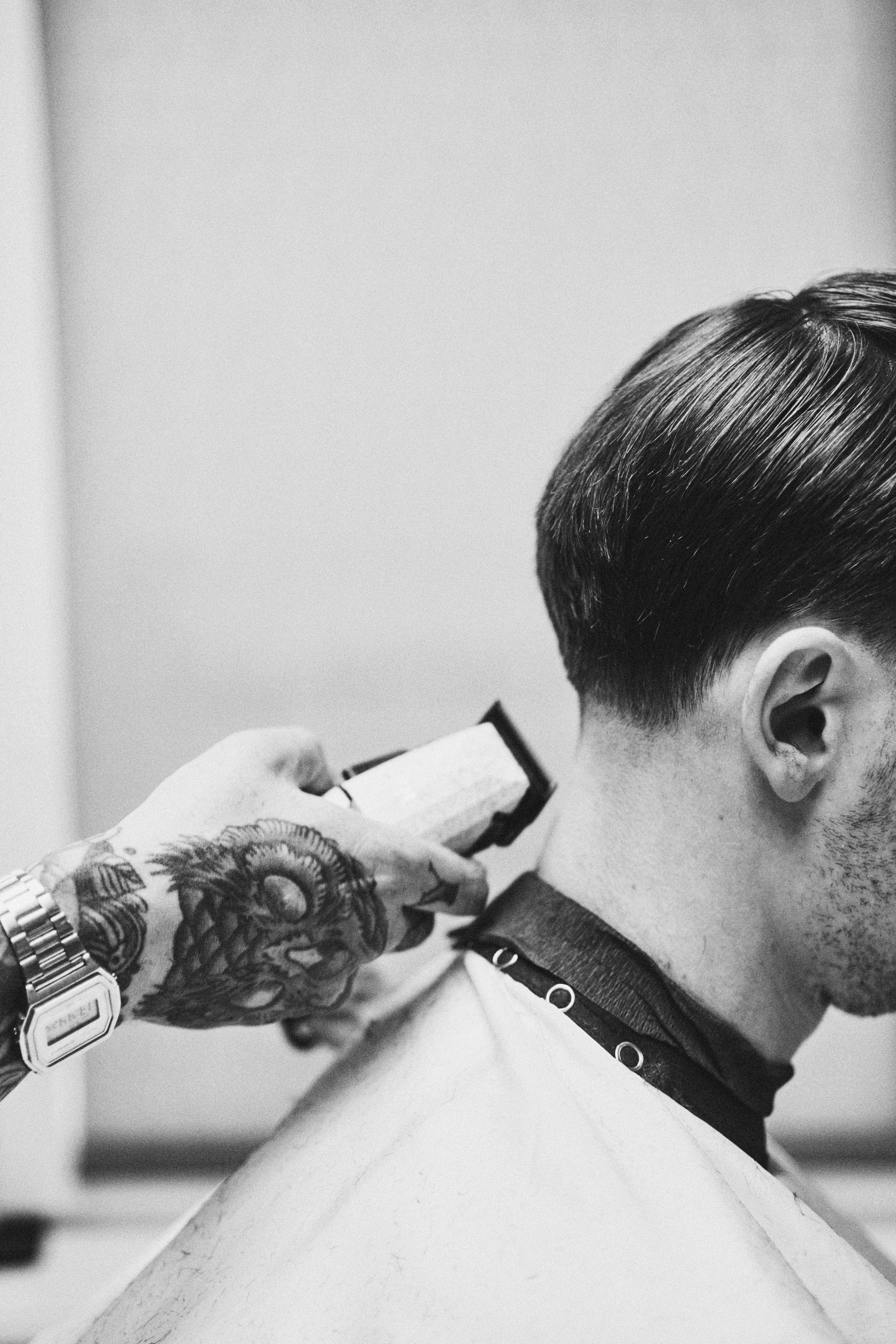 Black and white image of a barber with tattoos using a clipper on a client's hair.