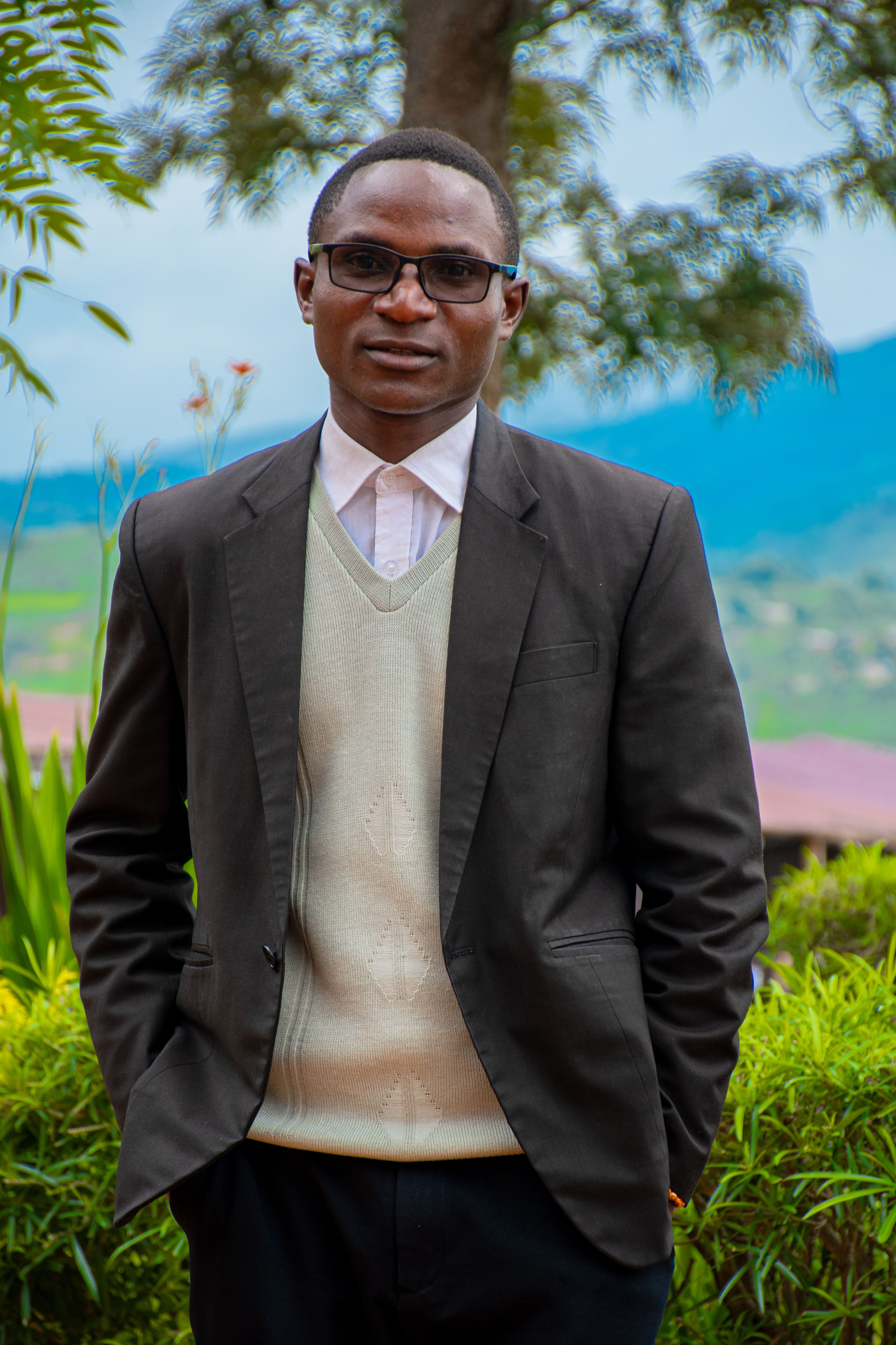 Man in formal attire standing outdoors with greenery and mountains in background.