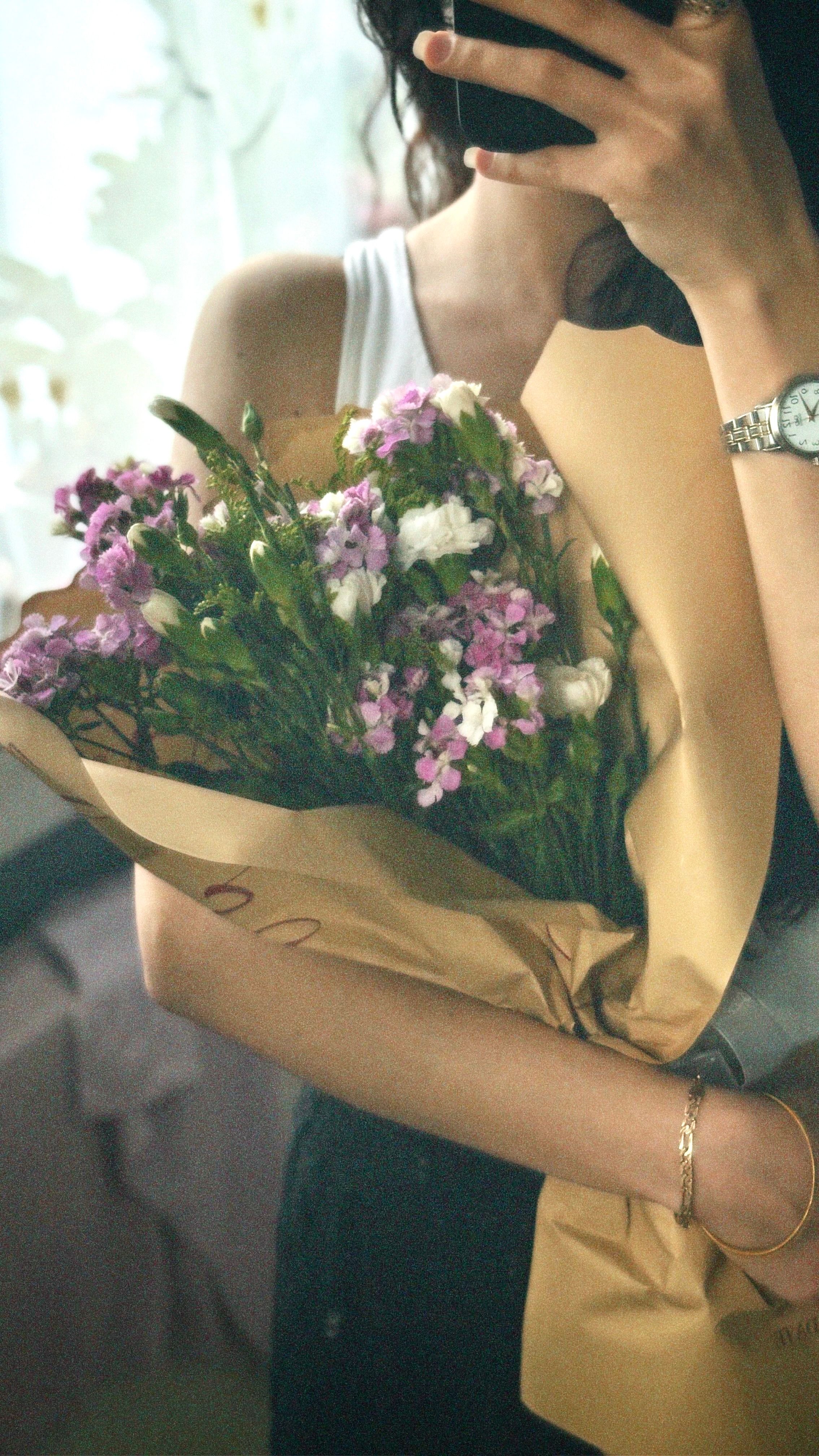 A woman holds a bouquet of purple and white flowers wrapped in brown paper indoors.