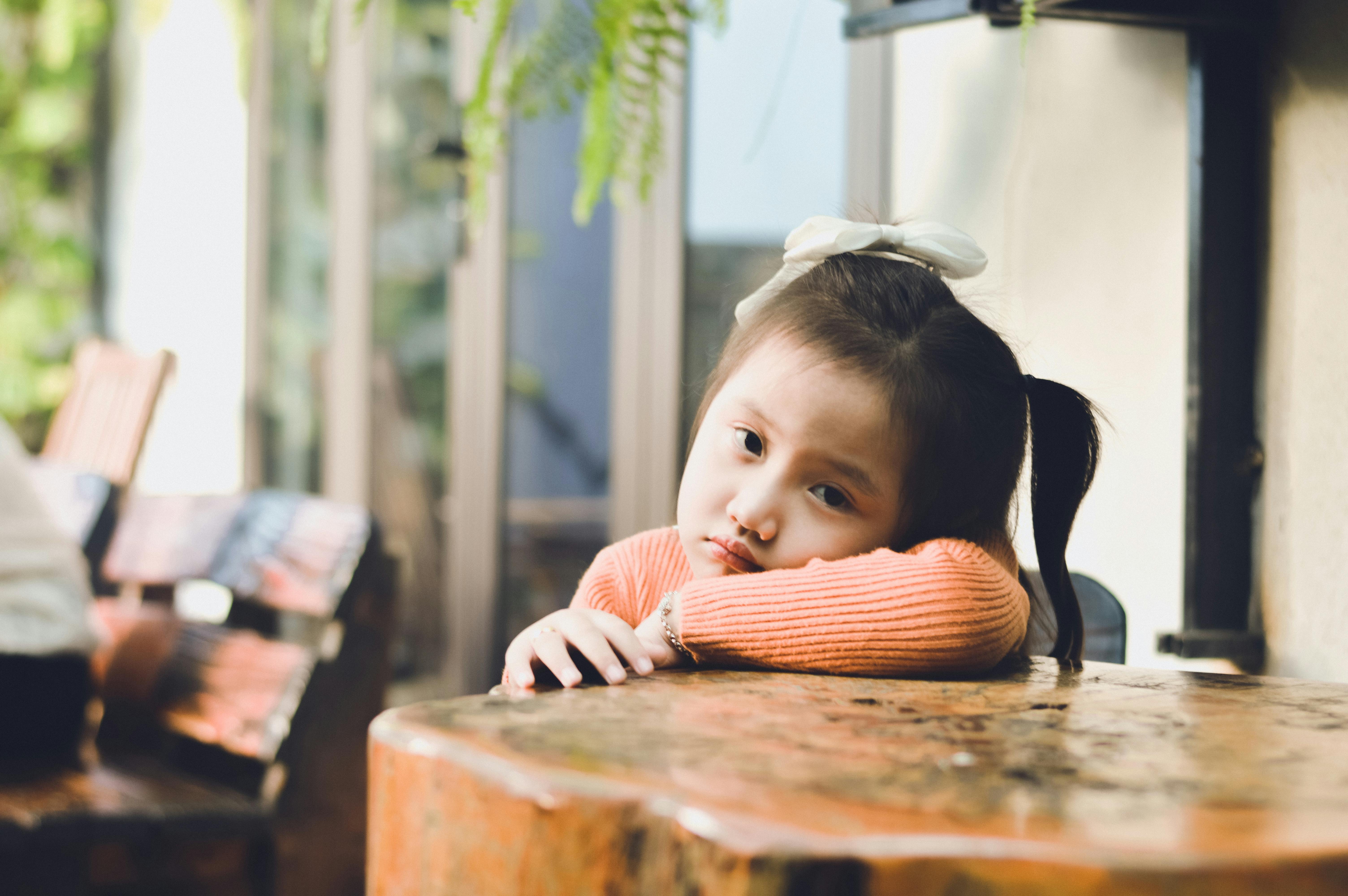A young girl, wearing an orange sweater, rests her head on a wooden table with a contemplative look.