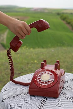 A hand lifting a red rotary phone receiver, set against a lush green field background.