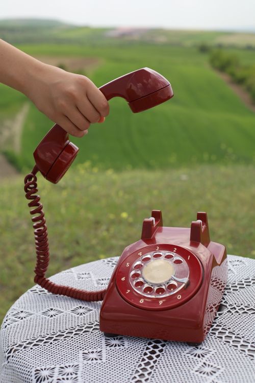Free A hand lifting a red rotary phone receiver, set against a lush green field background. Stock Photo