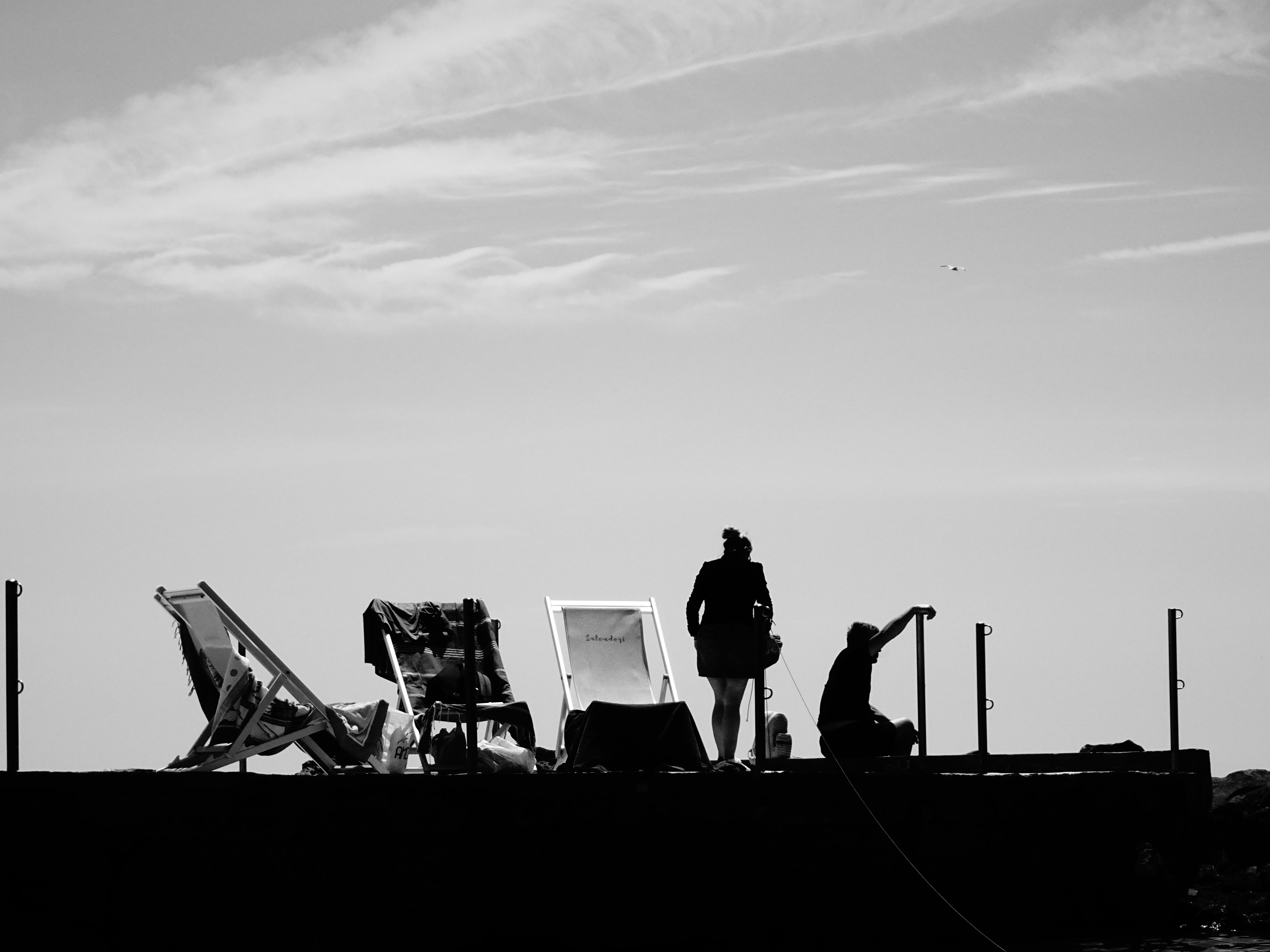 Gratis Silhouette in bianco e nero di persone su un molo con sedie a sdraio, che creano un'atmosfera serena e contemplativa. Foto a disposizione