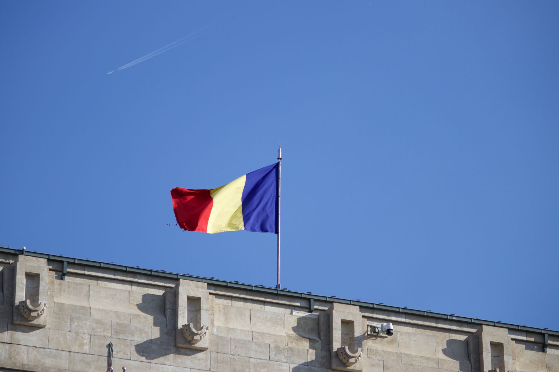 Romanian flag waving atop building under clear blue sky, aerial in background.
