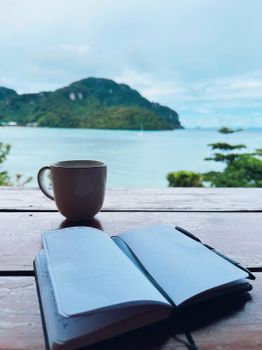 Open journal and coffee cup on a table by a serene beachside view.