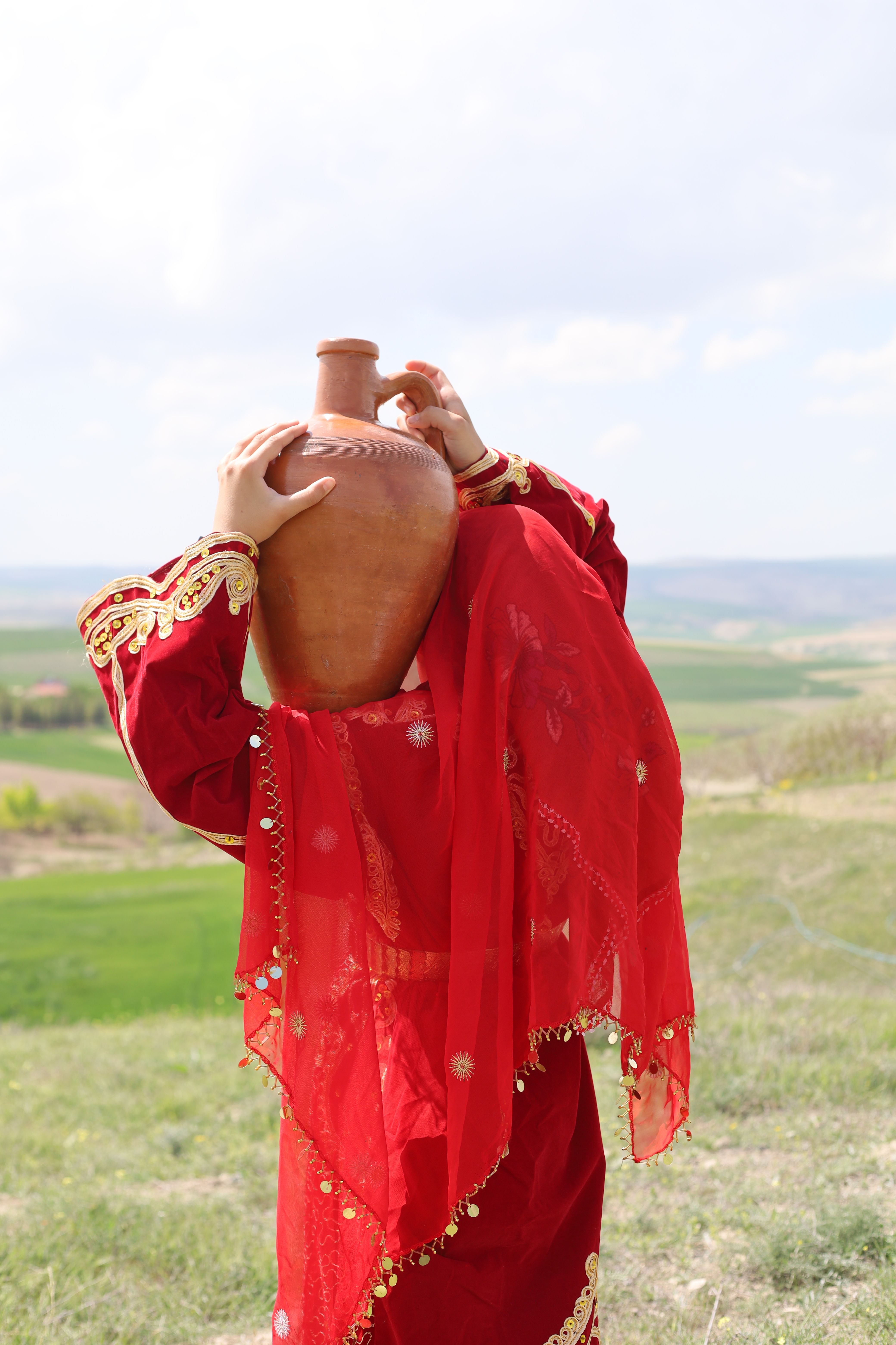 Woman in red traditional attire carries a clay pot outdoors, symbolizing cultural heritage.
