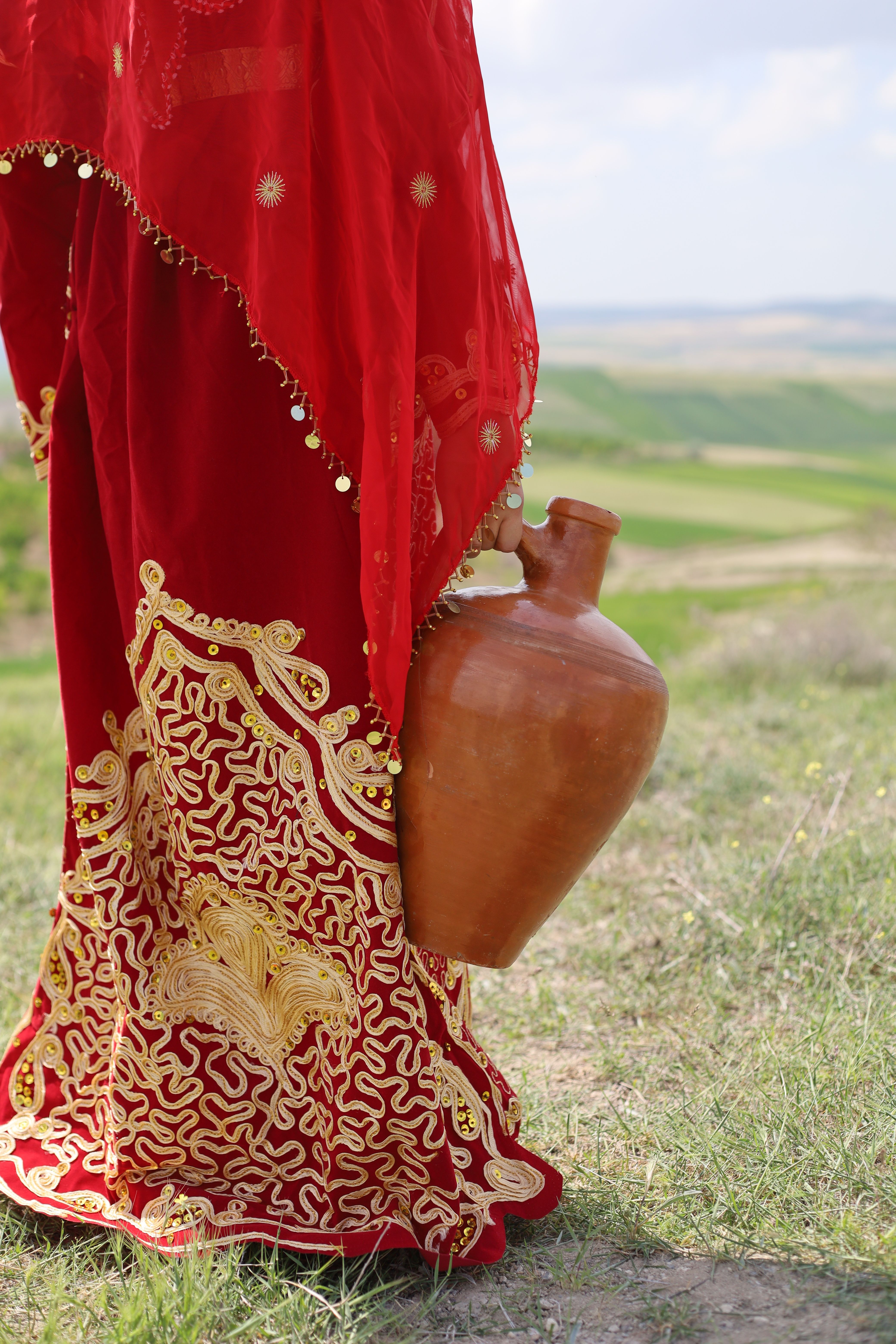 A person in ornate red clothing holds a clay jug in a serene countryside setting.