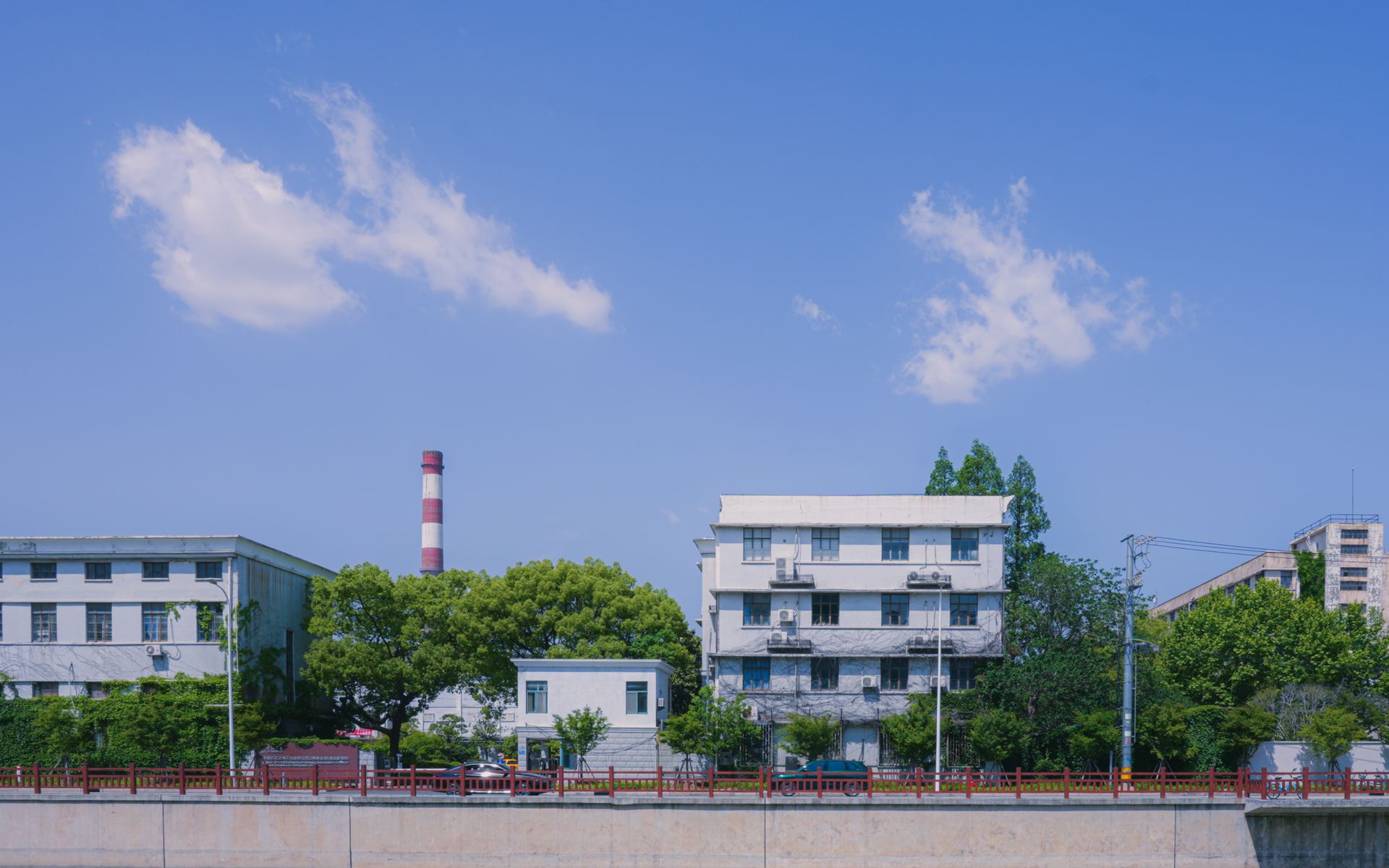 Industrial buildings and stack with trees under a clear blue sky, indicating a bright day.
