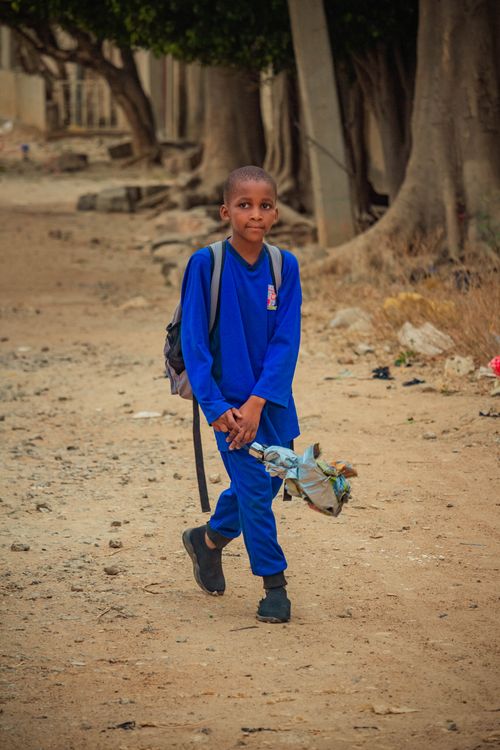 Free A young boy walking along a dusty path in a rural setting, carrying a backpack and wearing casual attire. Stock Photo