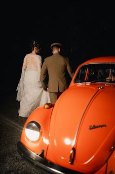 Bride and groom walk beside vintage orange car at night.