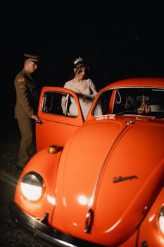 Bride in wedding dress enters vintage orange car assisted by uniformed man at night.