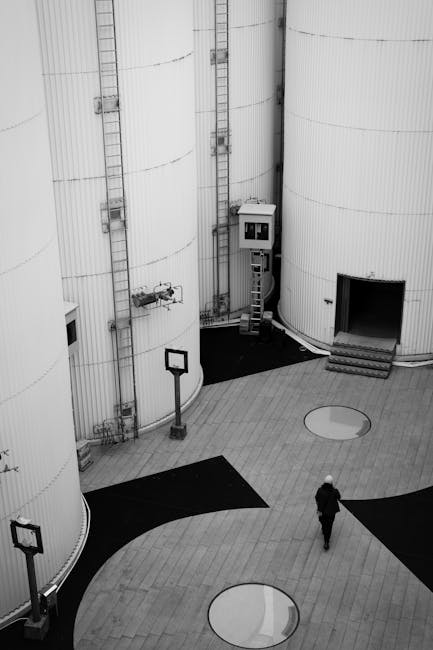 Black and white image of large industrial silos with a lone person walking below, highlighting scale and architecture.