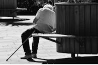 这是一张黑白照片，照片中一位拄着拐杖的年长男子坐在城市公园的长凳上