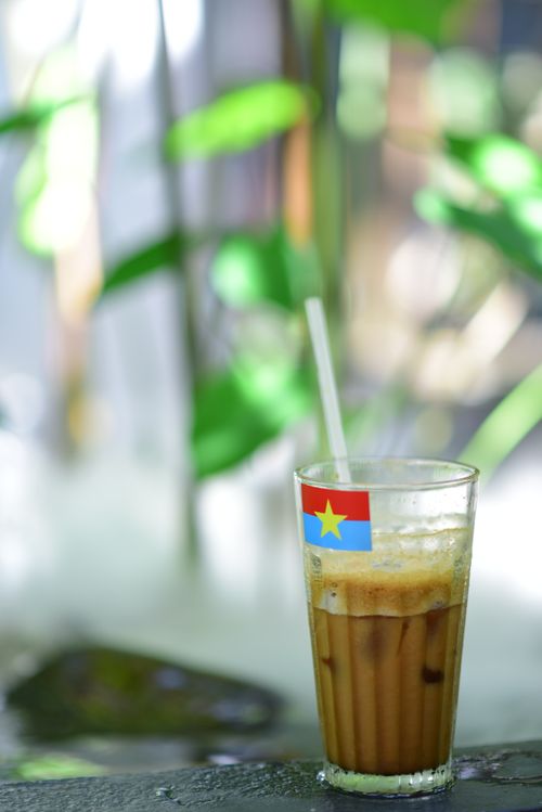 Free Glass of iced coffee with a flag sticker on a table surrounded by lush greenery. Stock Photo