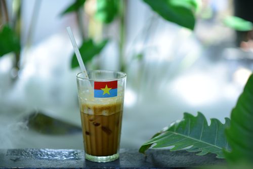 Free Glass of Vietnamese iced coffee with a flag design straw on a table surrounded by lush greenery. Stock Photo