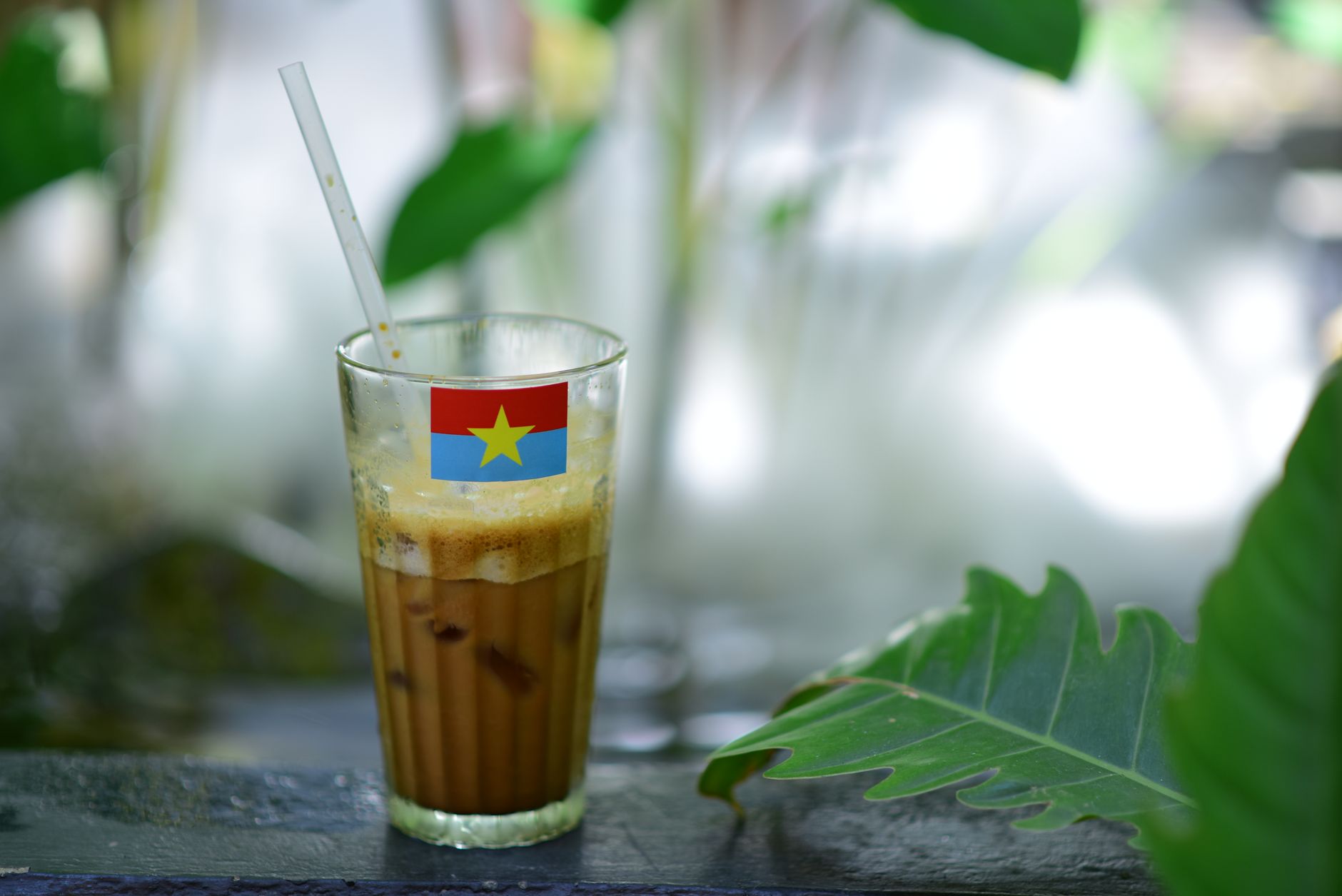 Glass of iced coffee adorned with Vietnamese flag outdoors with greenery.