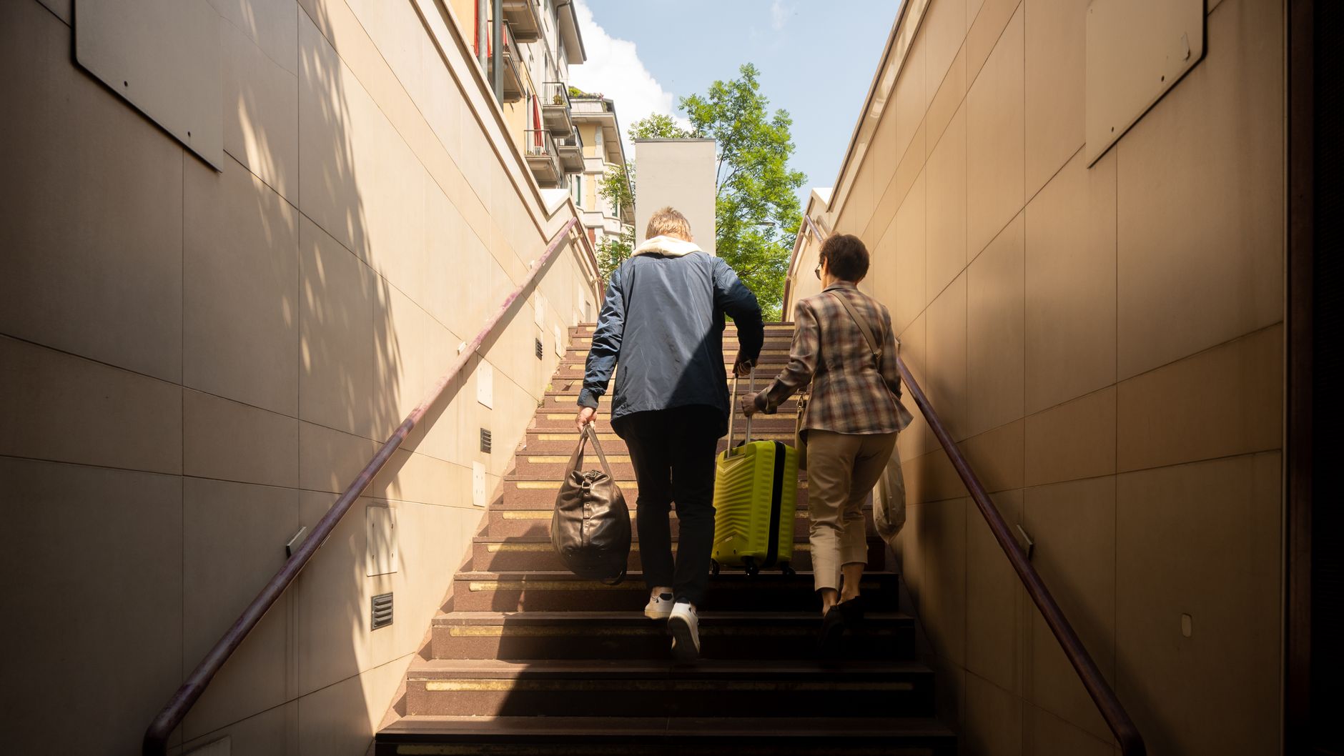 Back view of a couple carrying luggage up outdoor stairs on a sunny day.