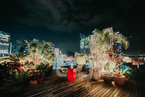 Rooftop with wooden floor and different potted plants near square table and stools with bright candle in front of contemporary buildings under serene sky at night