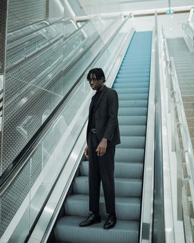 Fashionable young man in black outfit on an escalator indoors, showcasing style and modern architecture.