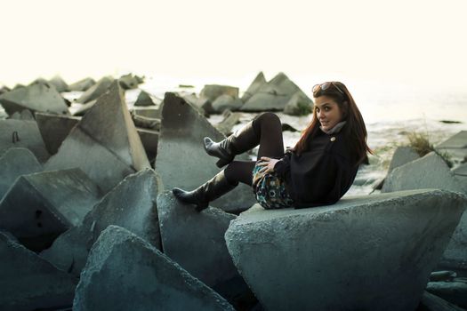 A fashionable woman in black boots posing on geometric concrete blocks near the sea.