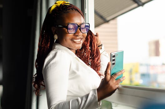 Happy woman in glasses and white blouse using smartphone by a window in Lusaka.