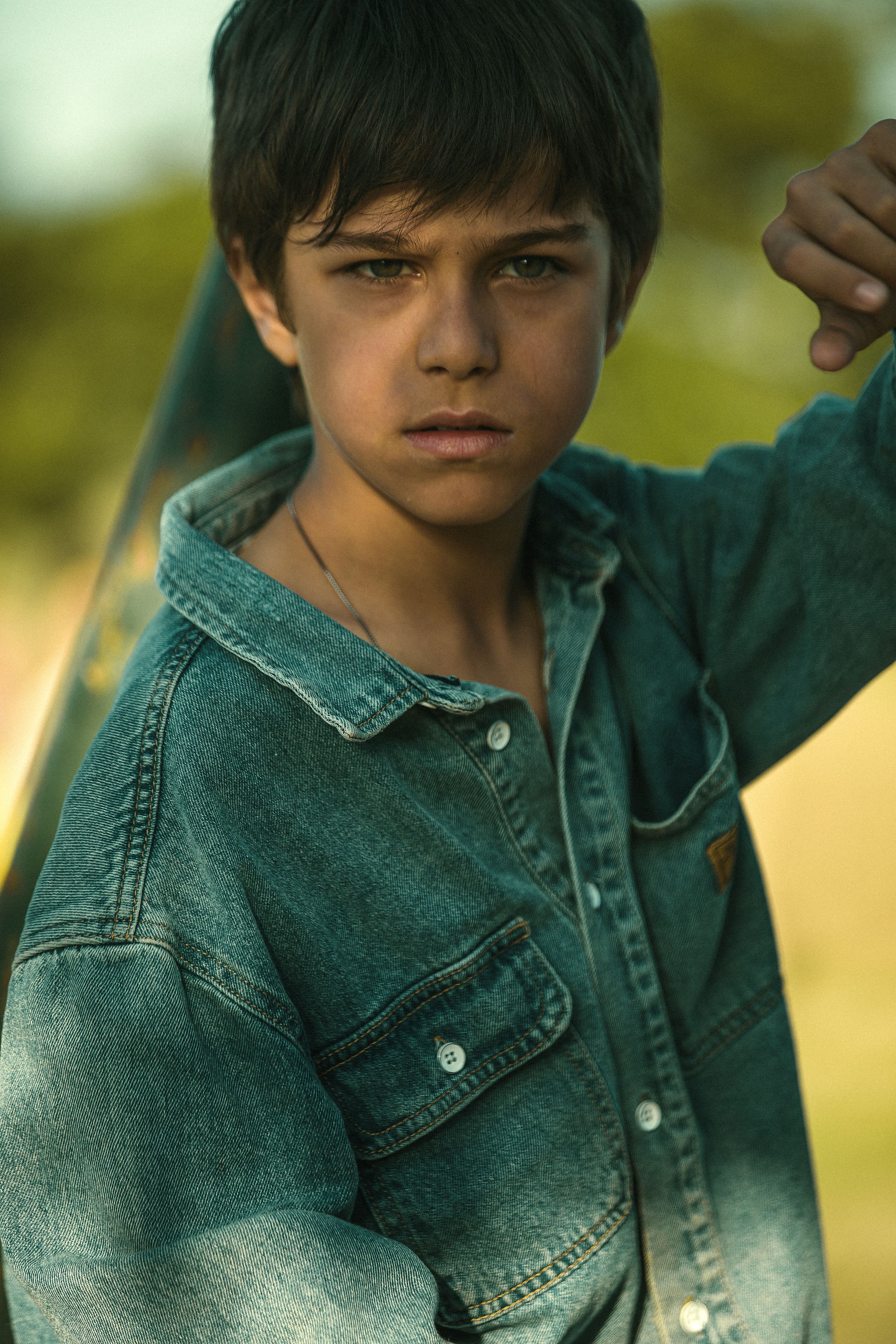 A serious young boy wearing a denim jacket poses outdoors, projecting confidence and determination.