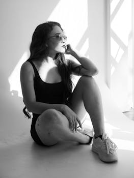 Black and white portrait of a woman sitting in sunlight, wearing glasses and casual outfit.