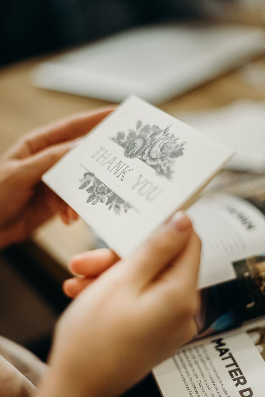 Close-up of hands holding a thank you card with floral design. Soft focus, warm indoor lighting.