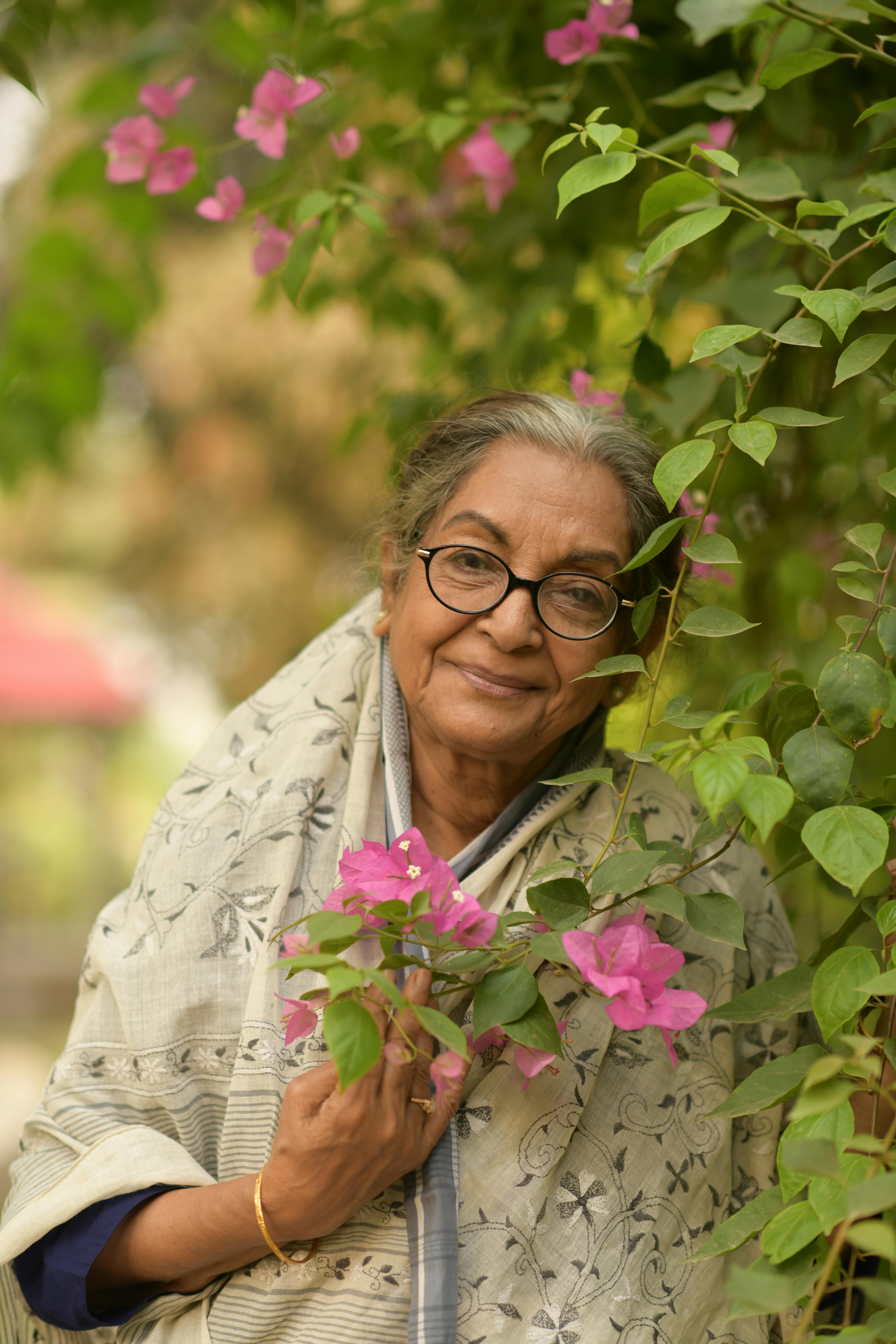 A senior woman smiling in a garden with vibrant Bougainvillea flowers.